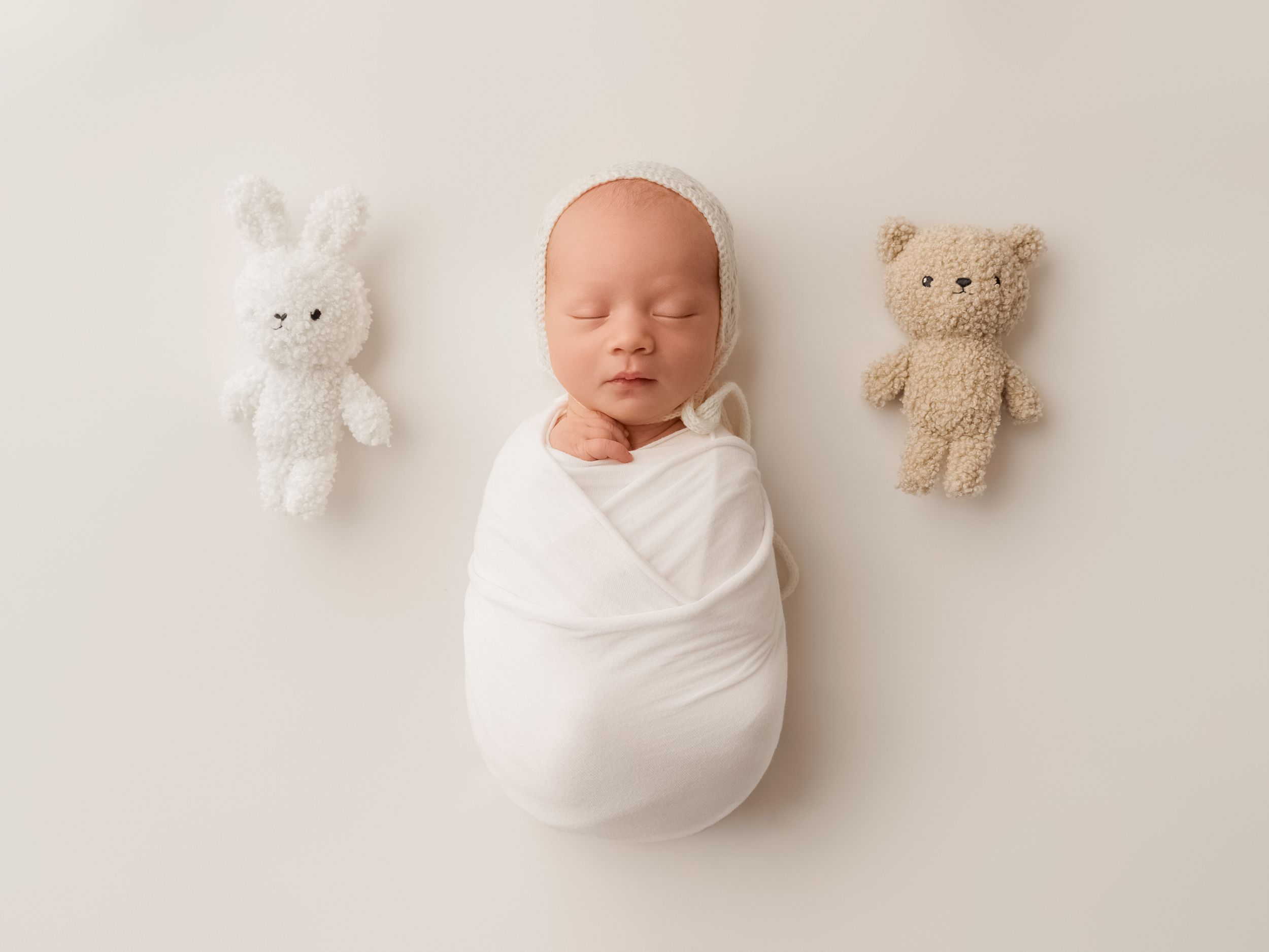 Newborn baby swaddled in white wrap with bonnet, posed between a white bunny and brown teddy bear plush toys.
