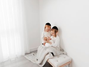 Two children in white outfits and black kippot sit on a bench, looking at a newborn baby wrapped in a striped blanket.