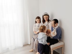 Family portrait with mother, two daughters, and baby in a bright white room near a curtained window.