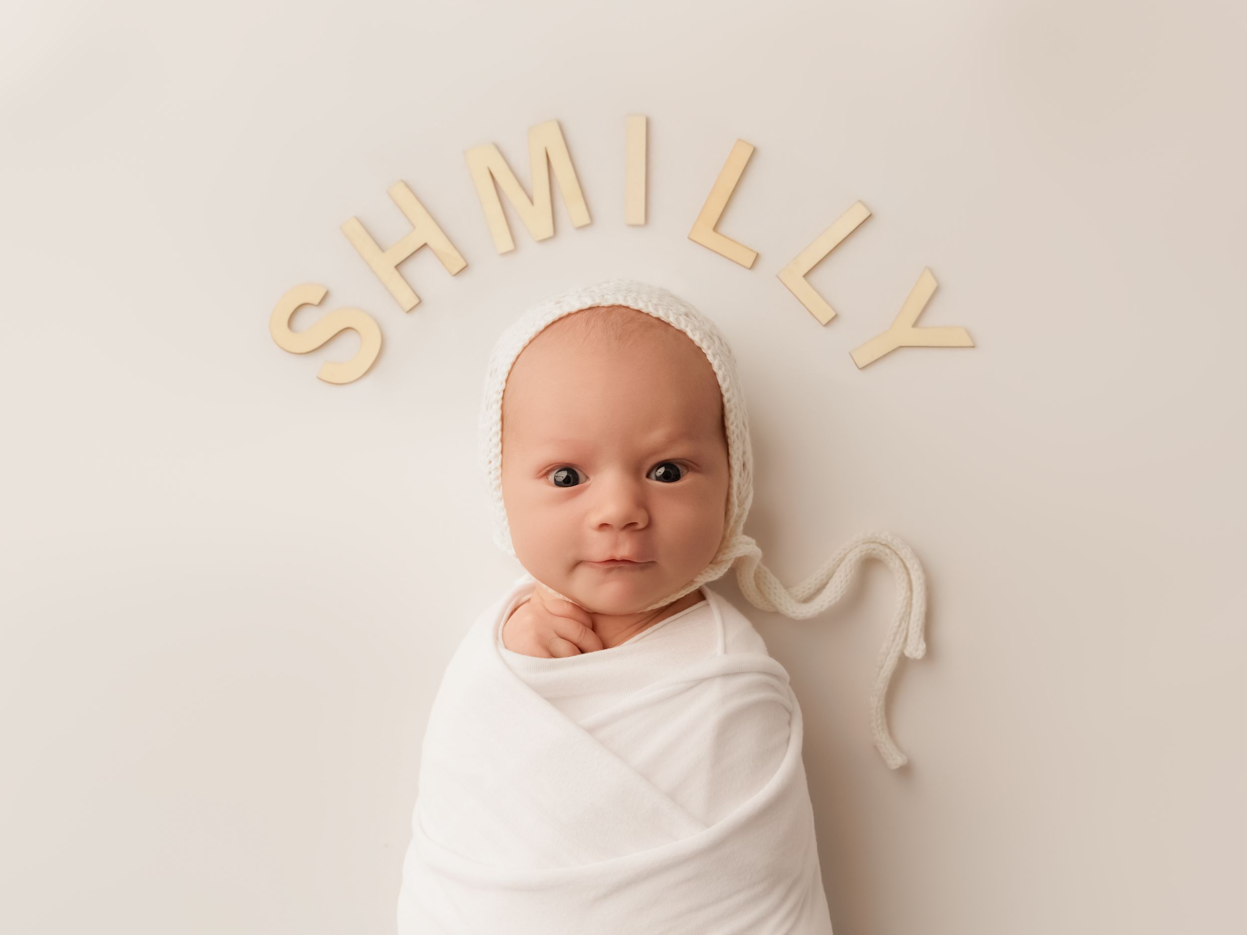 Newborn baby swaddled in white with wooden letters spelling SHMILLY above on a cream background.