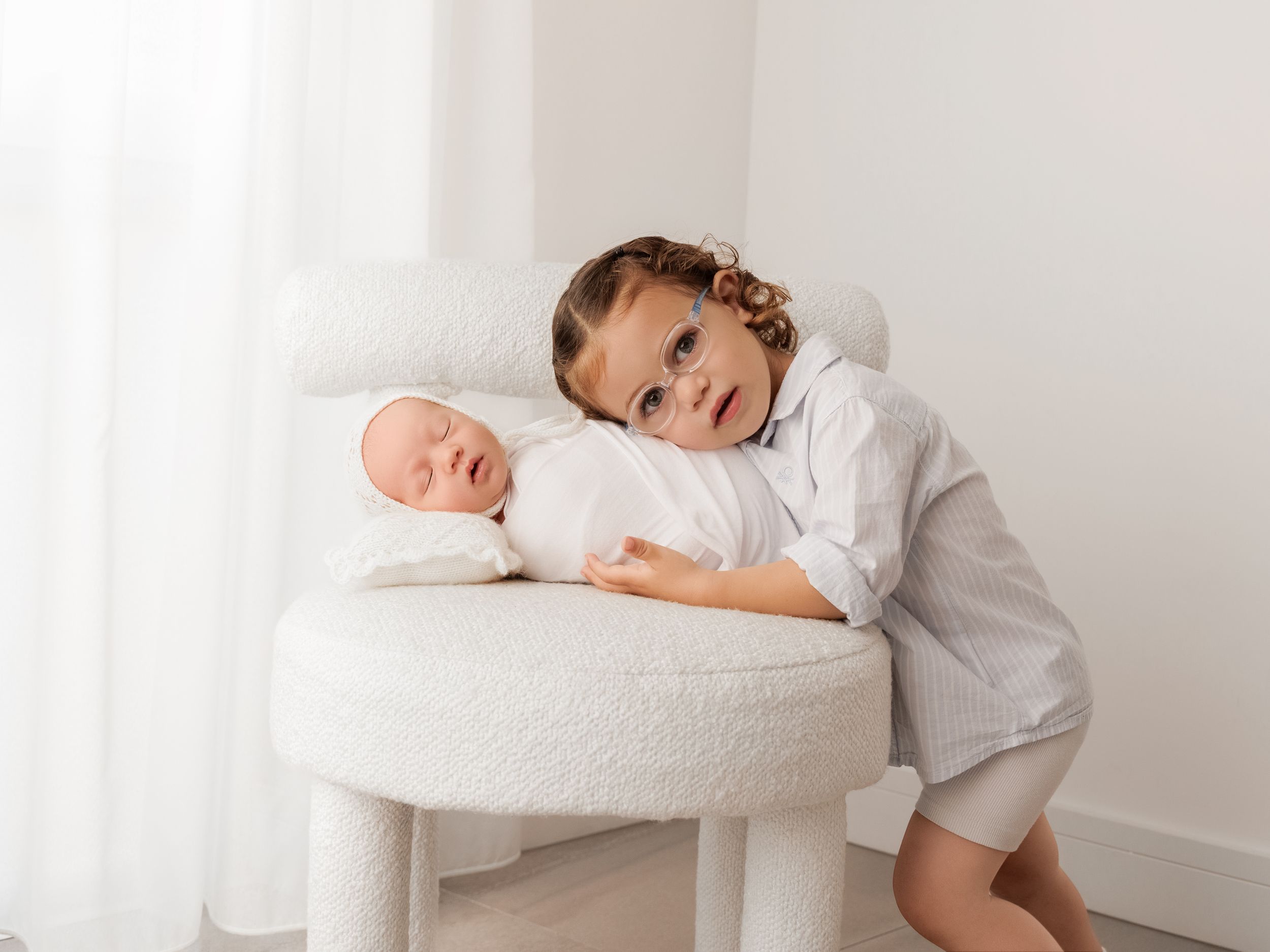 A toddler girl in white leans over a small round chair holding a swaddled newborn baby in a bright white studio.