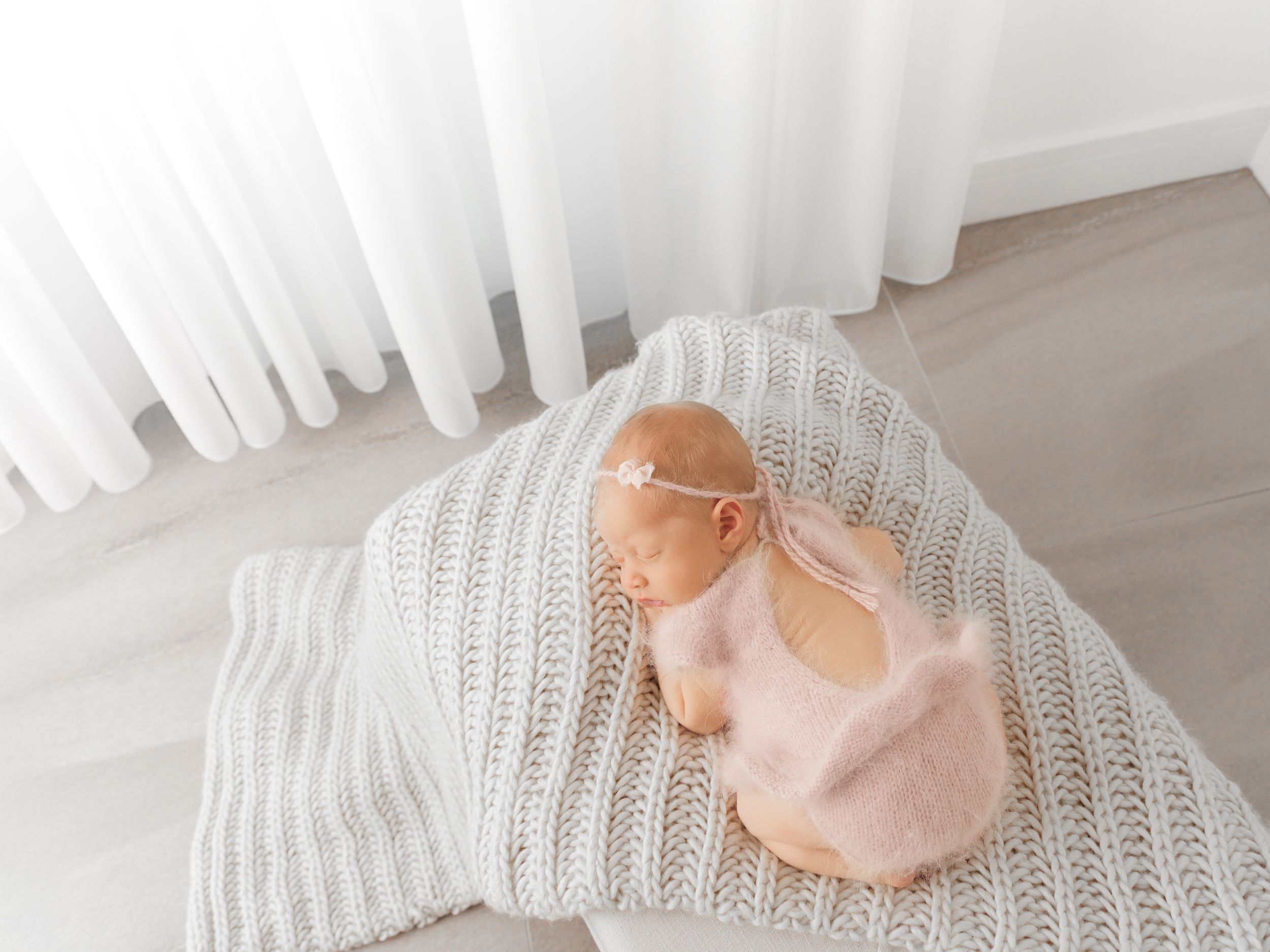 Newborn baby sleeping on a white knit blanket with a pink wrap and floral headband near a bright window.