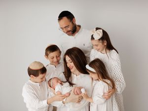 Jewish family in white outfits gathered around mother holding newborn baby in a bright studio portrait.