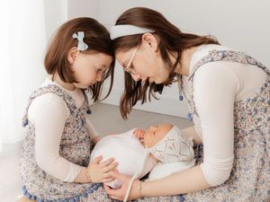 A mother and young girl in floral dresses lean over a sleeping newborn baby wrapped in white.