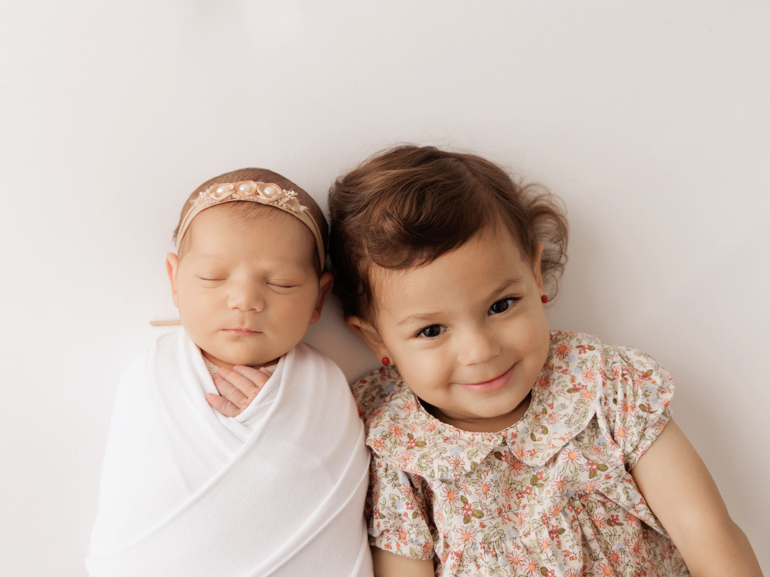 Newborn baby swaddled in white next to smiling toddler girl in floral dress on white background.