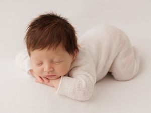 Peaceful newborn baby sleeping in white knit outfit with dark hair, posed on white background.