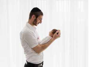 A proud father in a white shirt and kippah tenderly holds his swaddled newborn baby near a bright window.