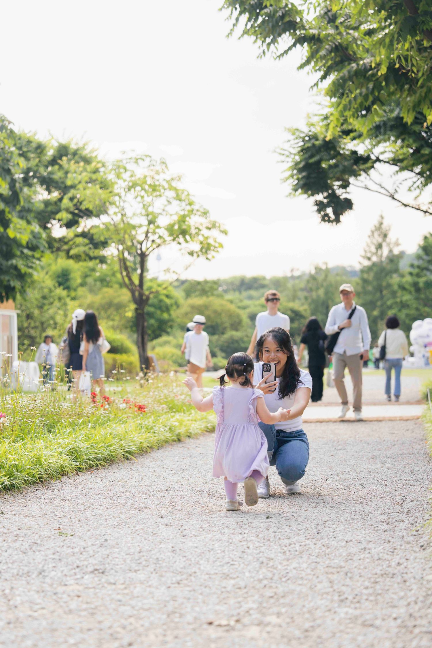 A mother crouches to take a selfie with her toddler on a scenic park path surrounded by lush greenery and flowers.