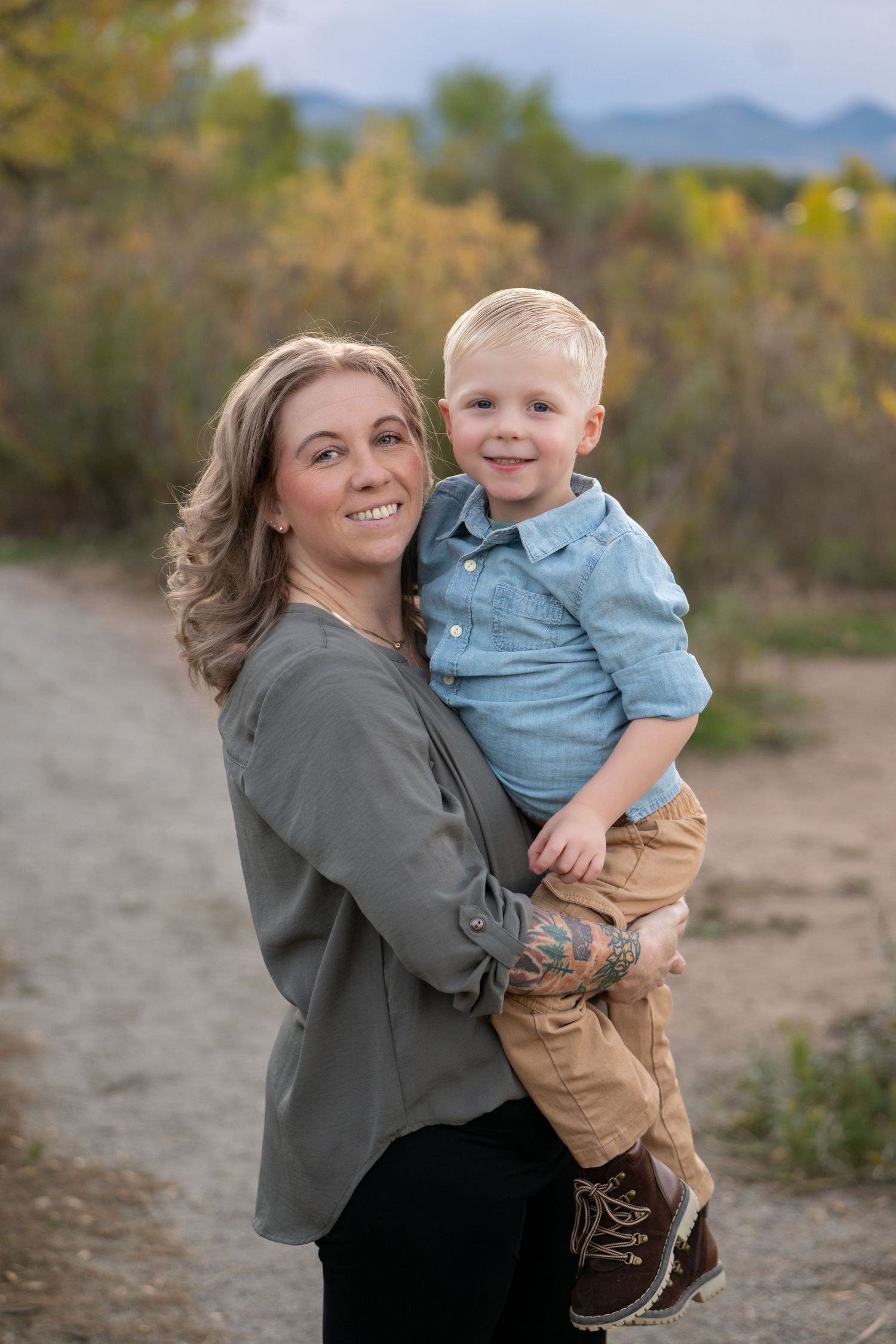 A smiling woman with tattoos holds a happy toddler boy outdoors on a dirt path with mountains in the background.