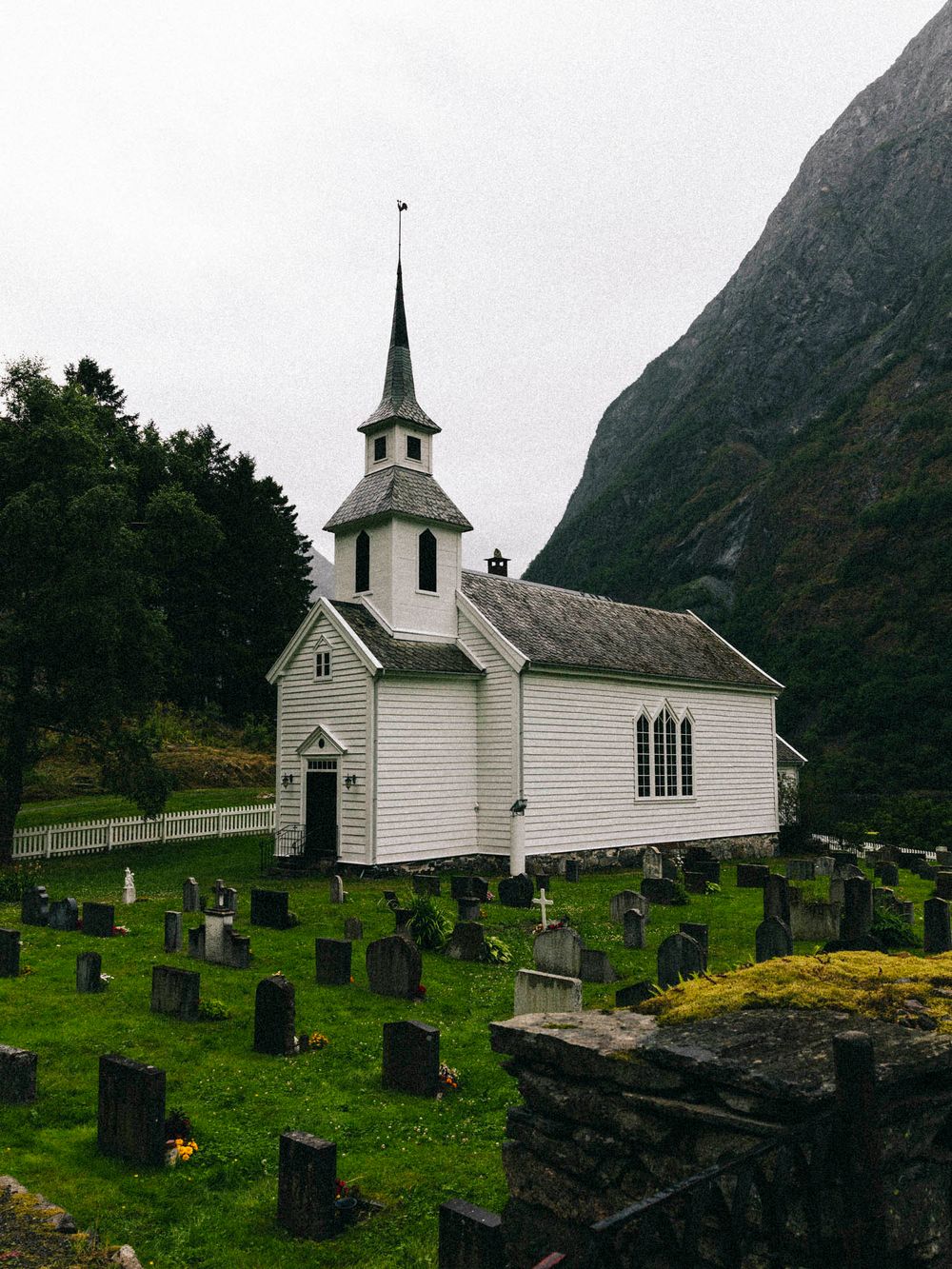 Historic white wooden Norwegian stave church with pointed steeple surrounded by a traditional cemetery against a rocky mountain.