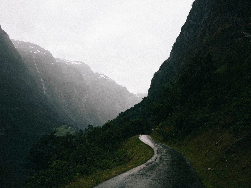 A winding road cuts through a dramatic misty mountain valley with steep rocky cliffs and lush green slopes.