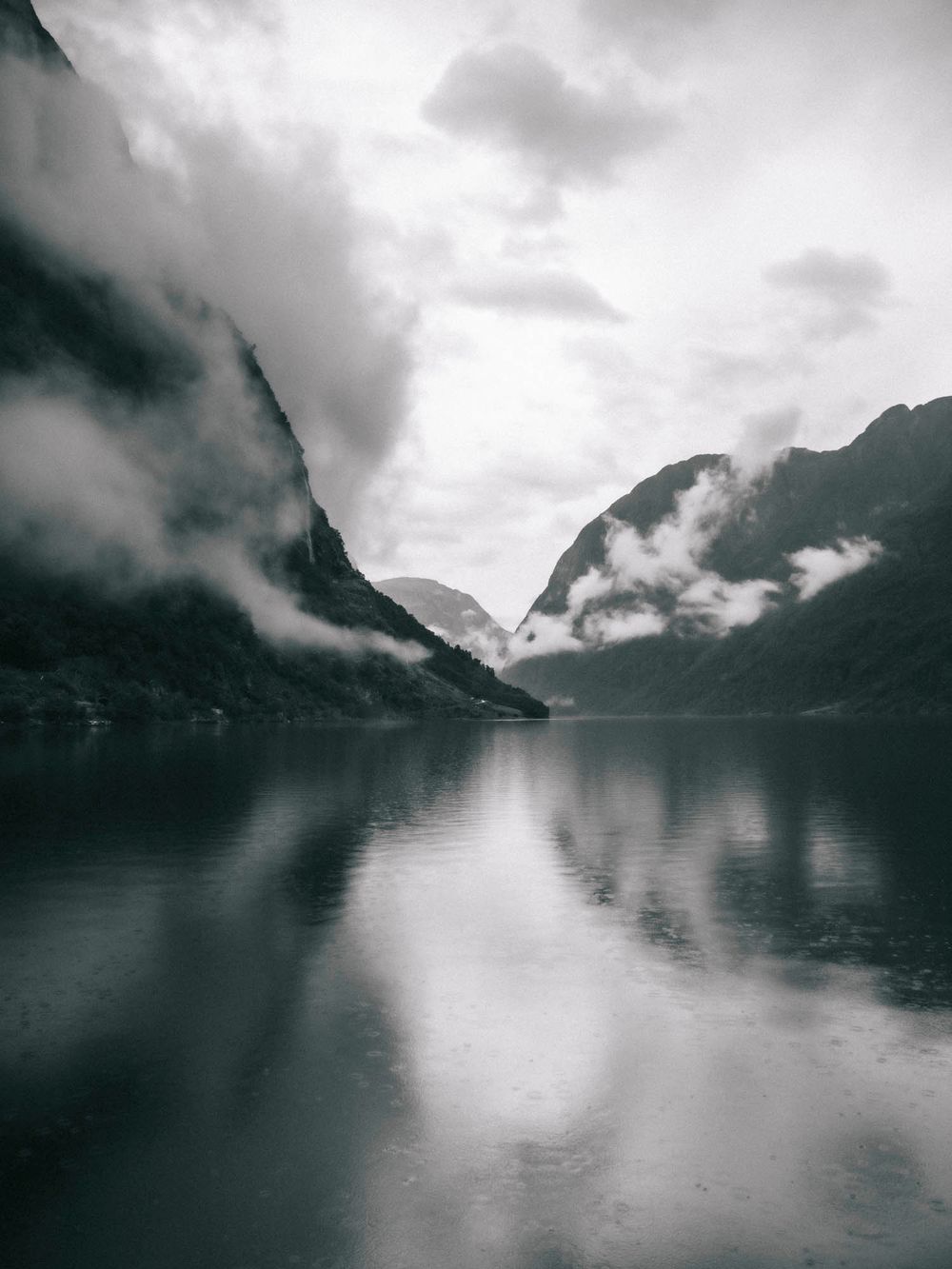 Dramatic black and white Norwegian fjord landscape with misty mountains reflected in calm water under cloudy skies.