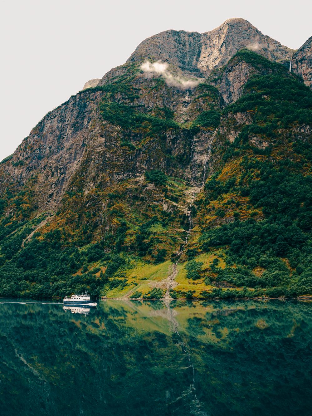 A white ferry boat glides across a calm fjord reflecting a dramatic green mountain with waterfalls in Norway.