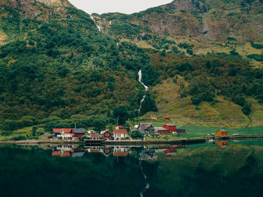 A serene Norwegian fjord village with colorful houses nestled at the base of a lush green mountain with a cascading waterfall.