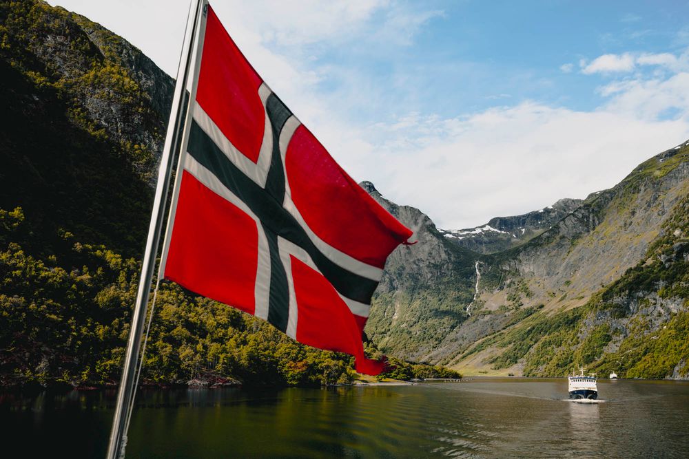 Norwegian flag waving over a scenic fjord surrounded by lush green mountains and calm waters with a boat in the distance.