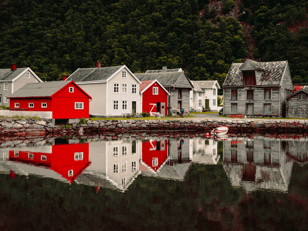 Red and gray wooden houses reflecting in calm fjord waters with lush green forested hills in Norway.