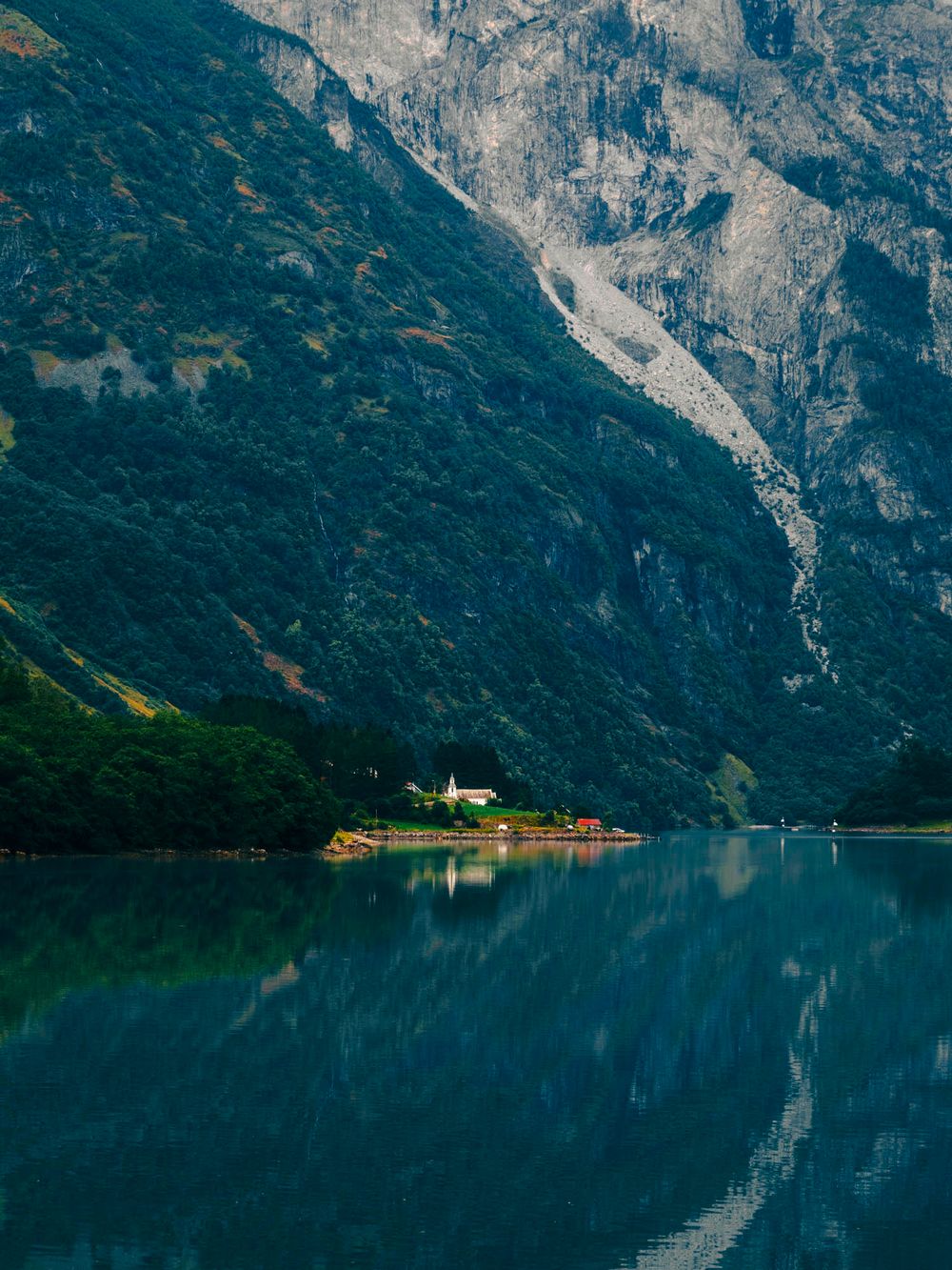 Serene Norwegian fjord with turquoise water reflecting lush green mountains and a small white building on the shoreline.