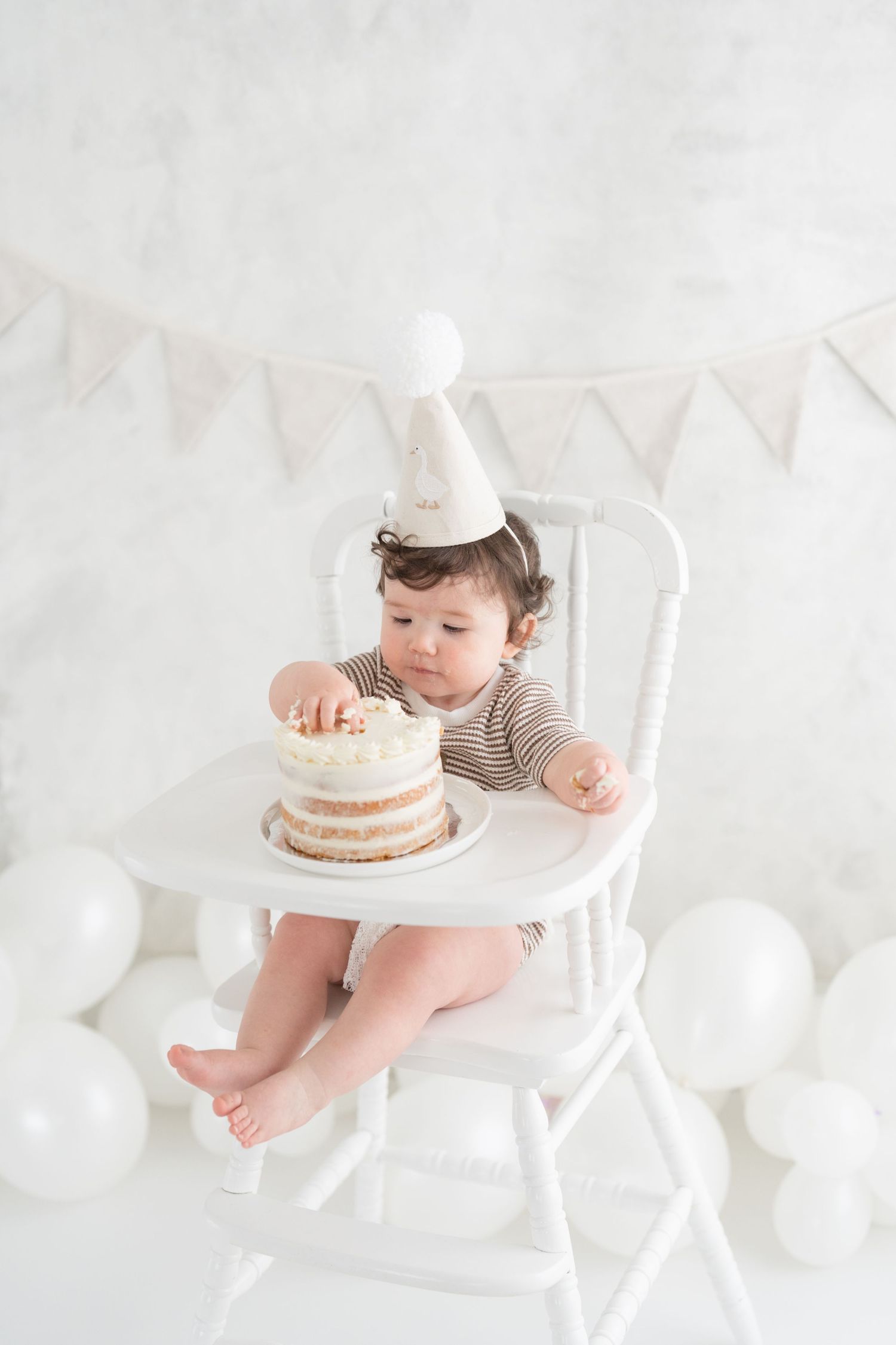 Bébé de 1 an assis sur une chaise haute antique dans un décor blanc et élégant pour une séance photo premier anniversaire avec gâteau