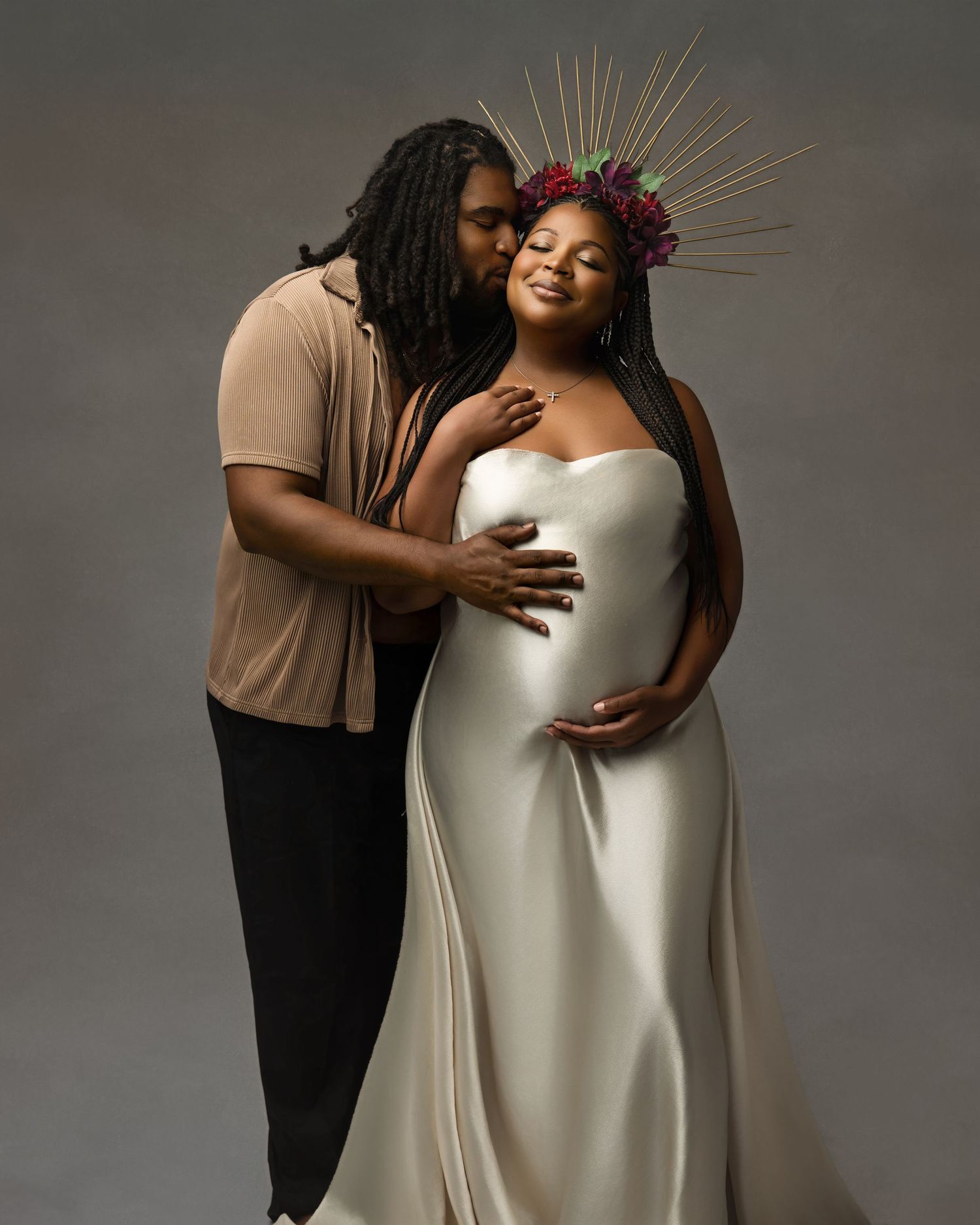Couple poses for maternity photo, woman in white gown with floral crown, partner kisses her cheek tenderly.