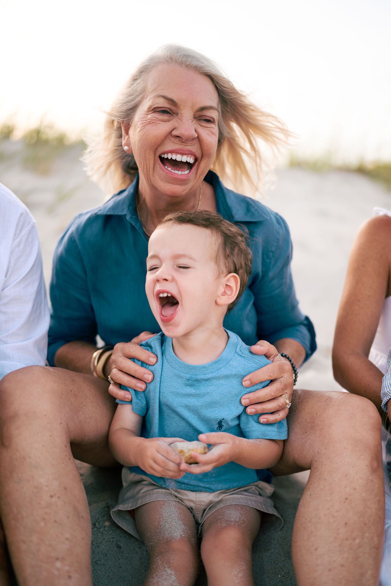 Grandmother and toddler grandson laughing joyfully together on a sandy beach during a sunny family outing.