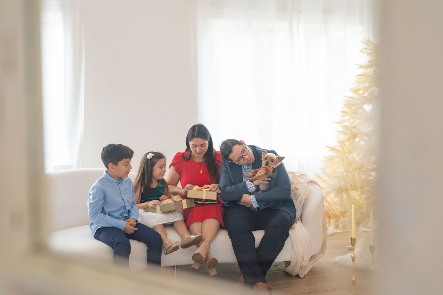 Family of five sitting on a couch exchanging Christmas gifts in a bright white holiday decorated living room.