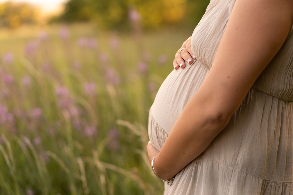 Pregnant woman in a flowing dress cradling her baby bump in a sunlit field of lavender flowers.