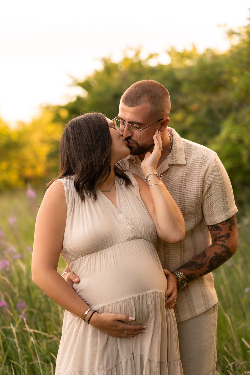 Pregnant woman in beige dress holds belly as tattooed partner kisses her cheek in golden hour field.