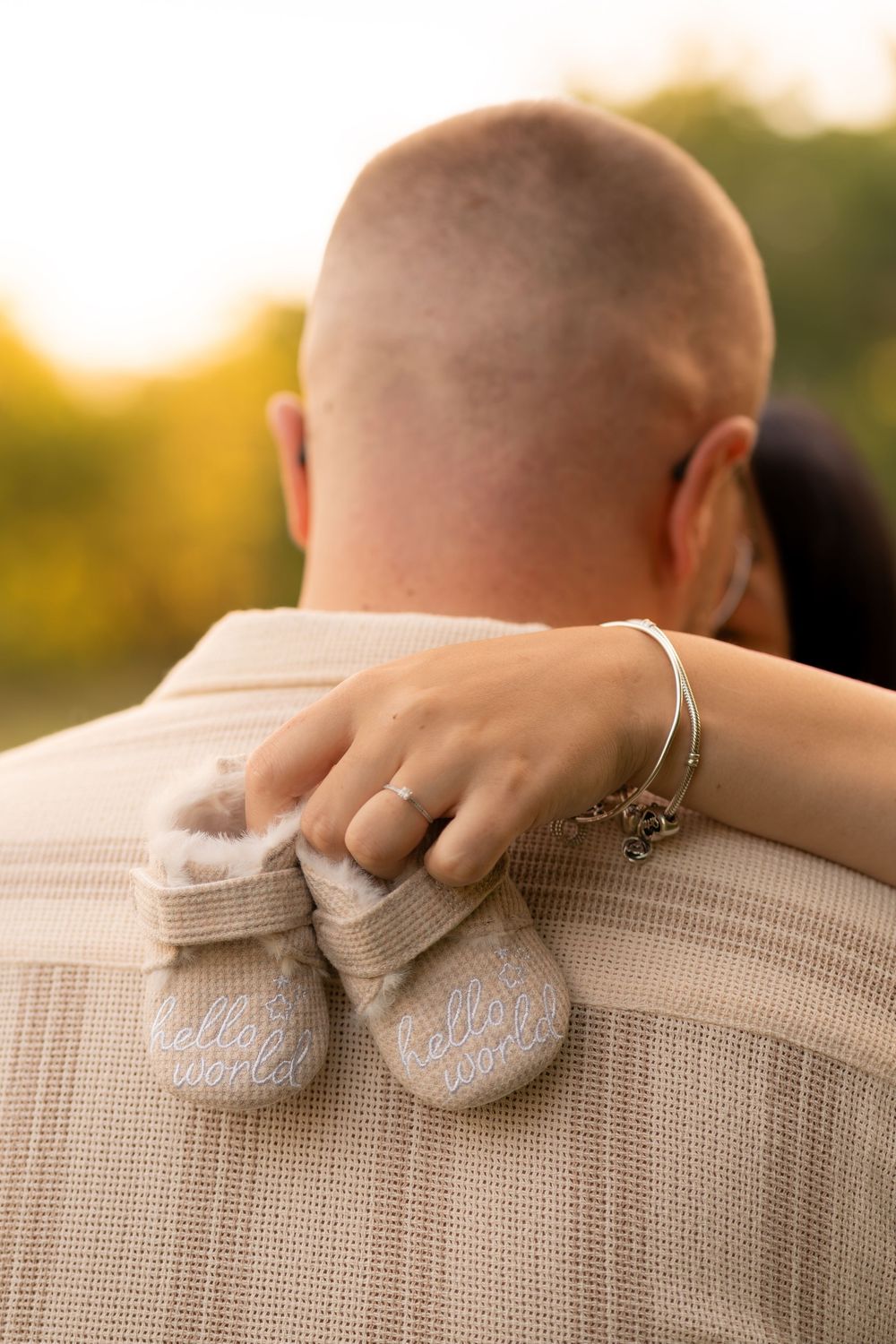 A woman rests her hand holding tiny baby booties on a bald man's shoulder during a golden hour maternity photo.