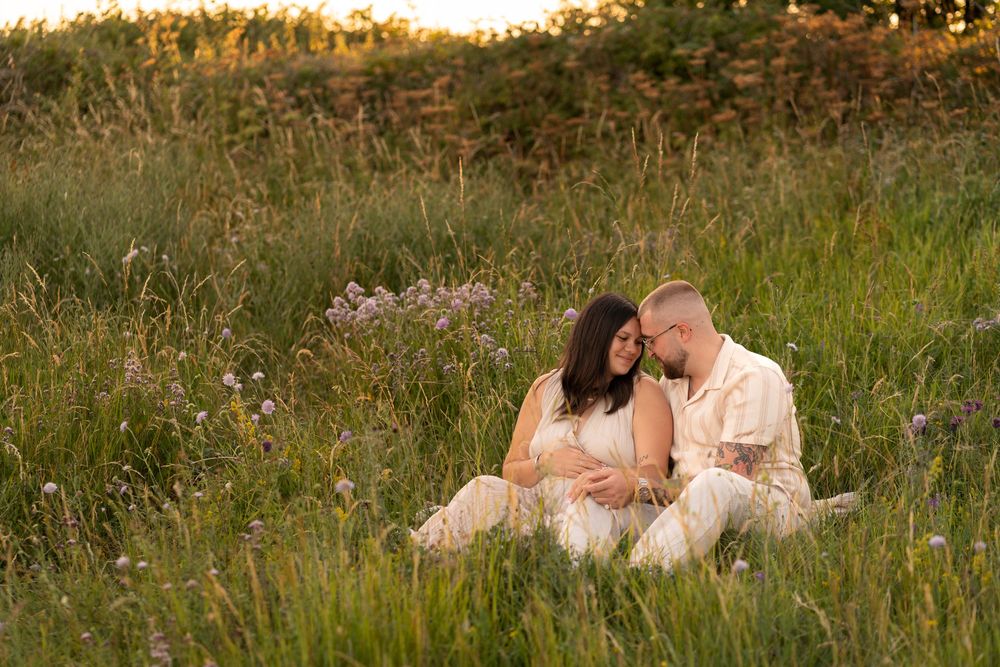 Couple sitting in a sunlit meadow, sharing a tender moment, with the woman visibly pregnant, surrounded by wildflowers.