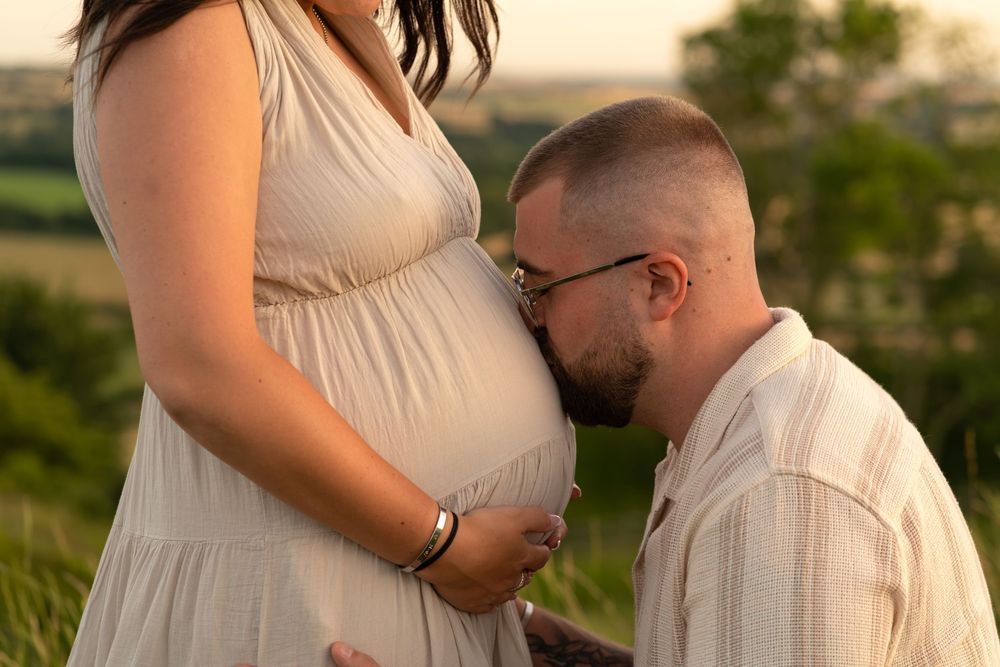 Man kneeling and kissing pregnant woman's belly outdoors during golden hour maternity photoshoot.