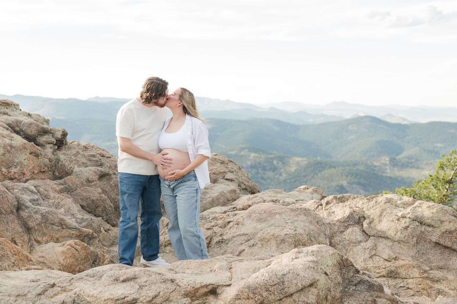 Couple shares a tender kiss on rocky mountain overlook, man touching pregnant woman's belly with scenic valley views behind them.