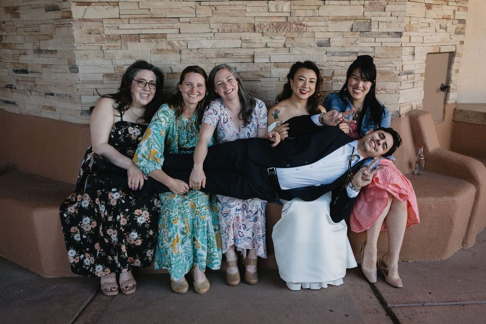 Joyful wedding party poses with groom being lifted by bridesmaids in colorful floral dresses.