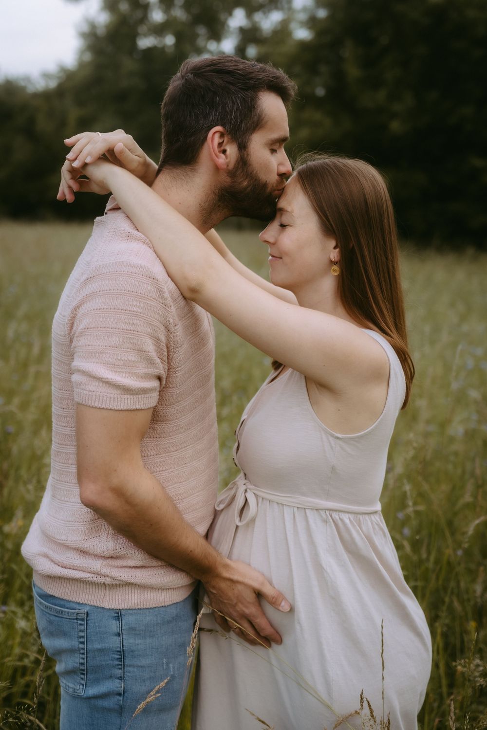 A couple shares a tender moment in a green field, woman in white dress embracing man in pink shirt.
