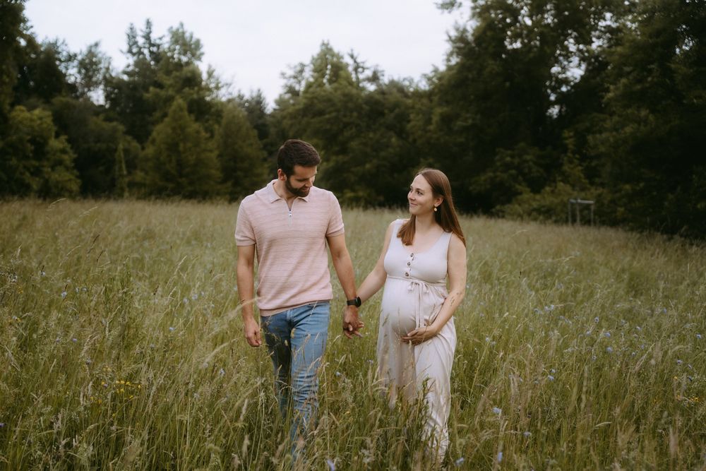 Pregnant couple holding hands walking through tall green meadow grass with trees in background.