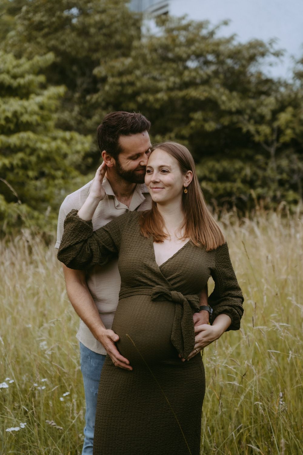 Happy couple embracing in tall grass field, pregnant woman in olive wrap dress with partner behind her.
