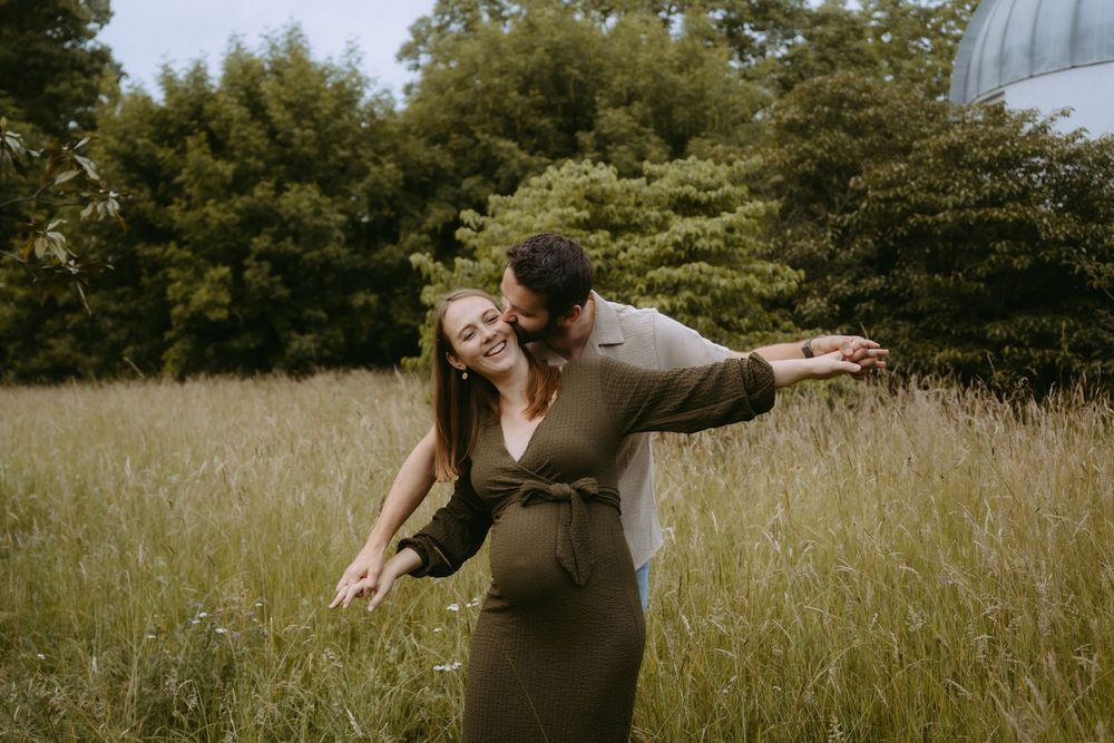 A couple embraces in a meadow, the woman in an olive dress with arms outstretched, as the man kisses her cheek.