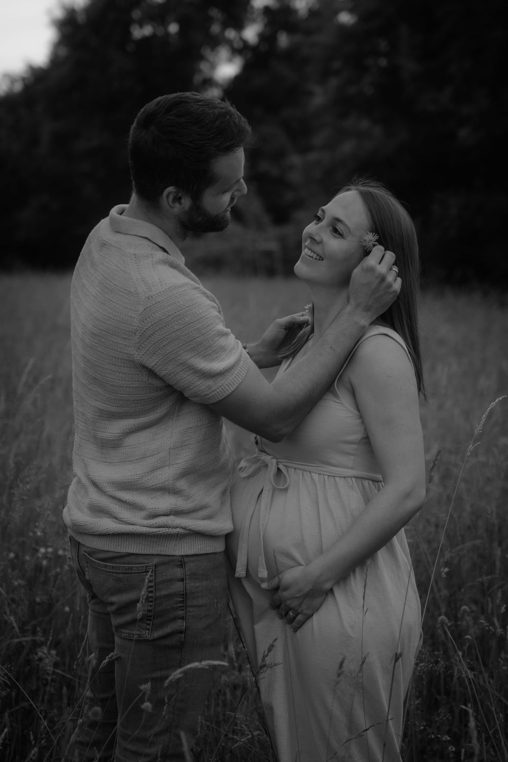 Black and white photo of a couple in a field, man tenderly brushing woman's hair as she smiles, visibly pregnant.
