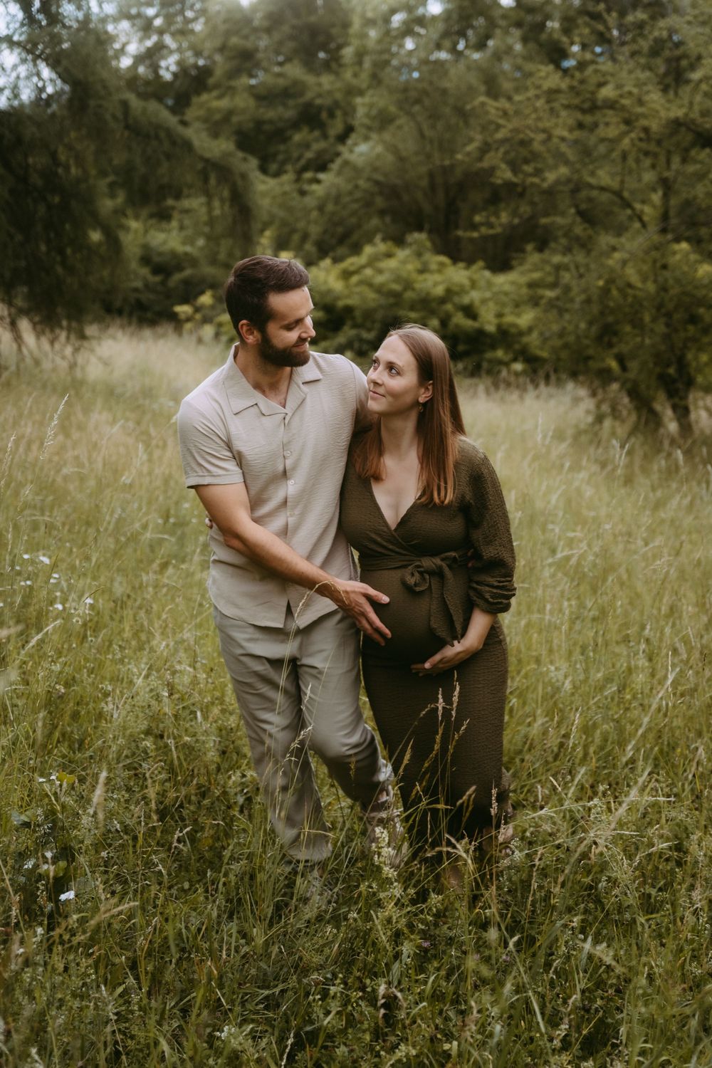 A couple stands in a lush green meadow, the woman in an olive dress cradling her baby bump as her partner embraces her.