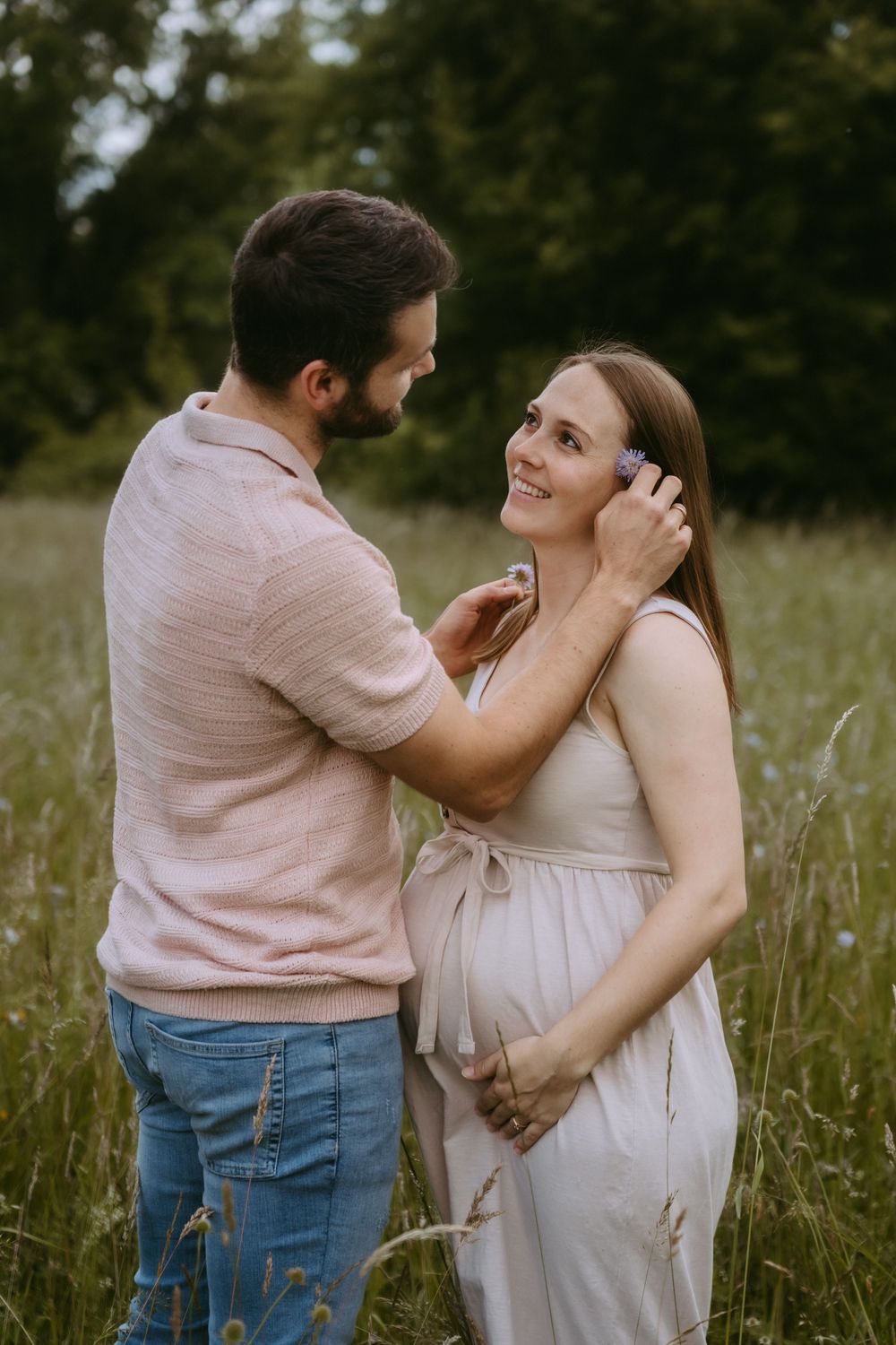 Smiling pregnant woman and bearded man sharing a tender moment in a lush green meadow field.