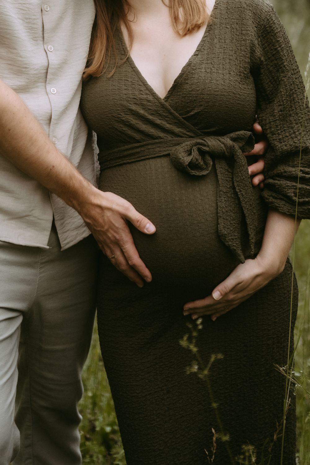 Couple sharing a tender maternity moment outdoors, man gently touching pregnant womans belly in olive wrap dress.