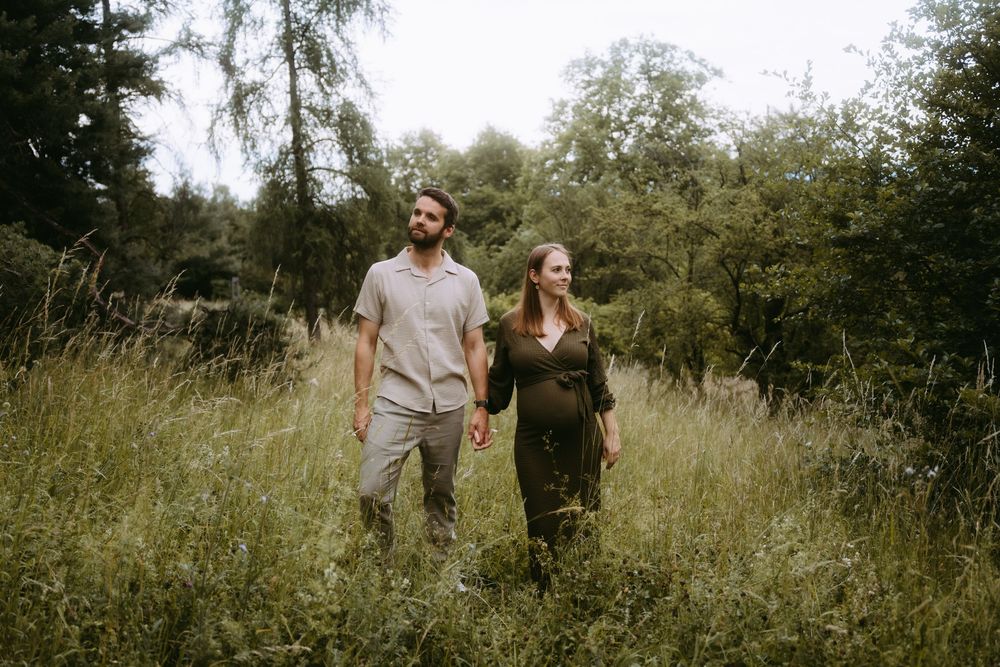 Expecting couple walking hand-in-hand through a lush green meadow surrounded by trees.