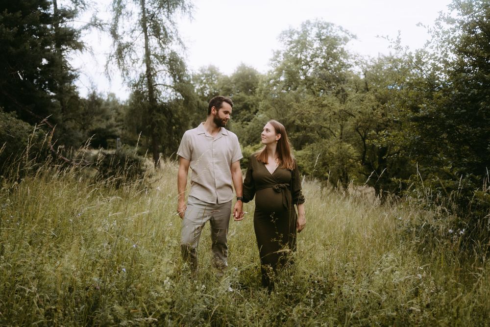 Couple holding hands walking through a lush green meadow surrounded by tall grass and trees.