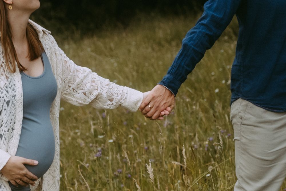 Pregnant woman holding hands with partner in a wildflower meadow, wearing a blue dress and white lace cardigan.