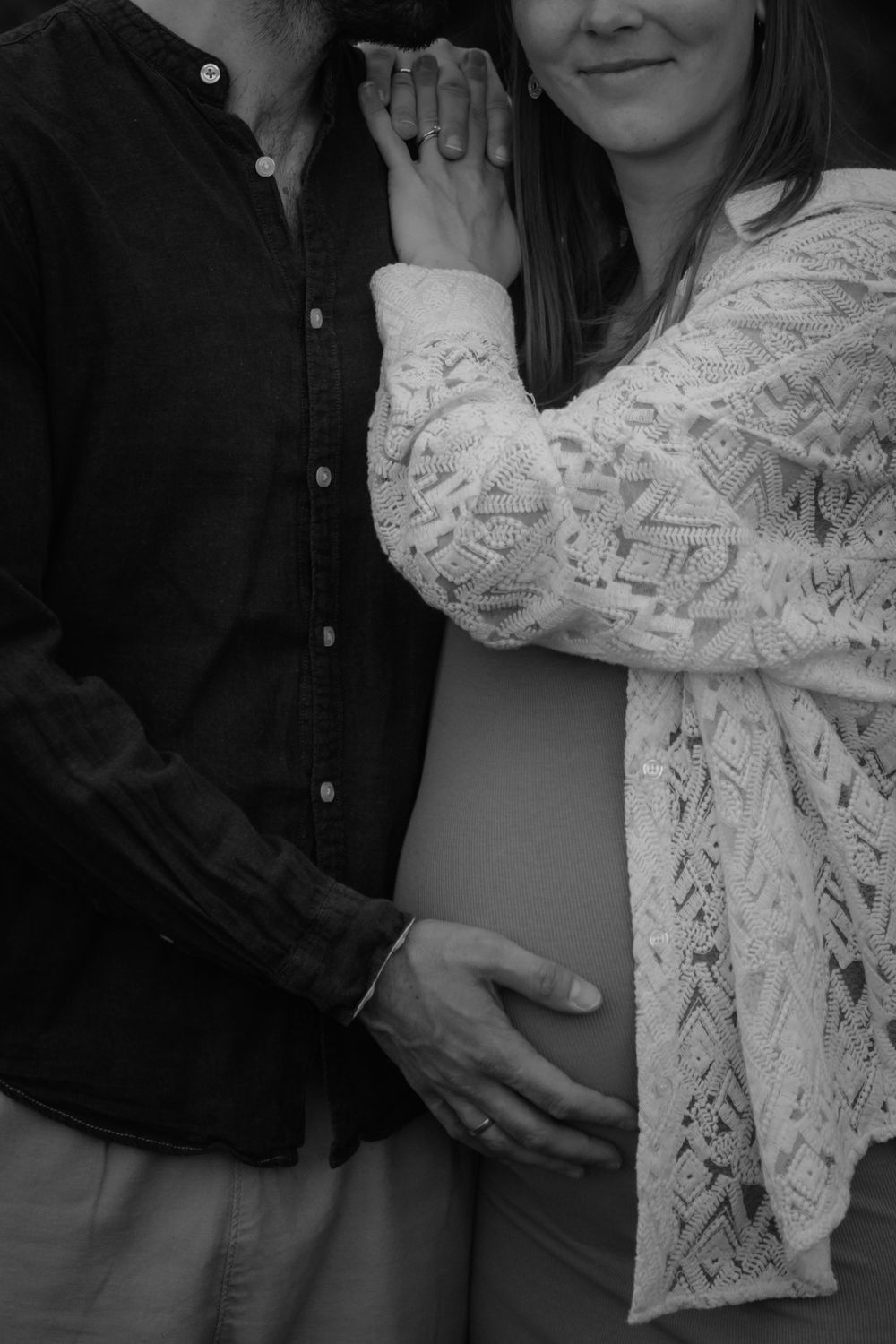 Black and white maternity photo of a couple holding hands and cradling a pregnant belly, wearing lace and dark shirt.