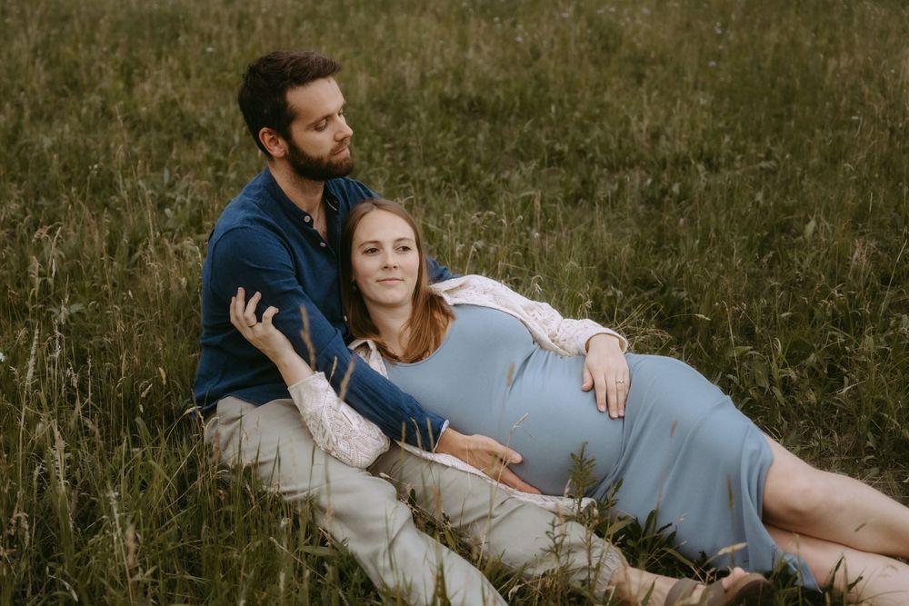 Pregnant woman in blue dress reclines against partner in grassy field during outdoor maternity photo session.