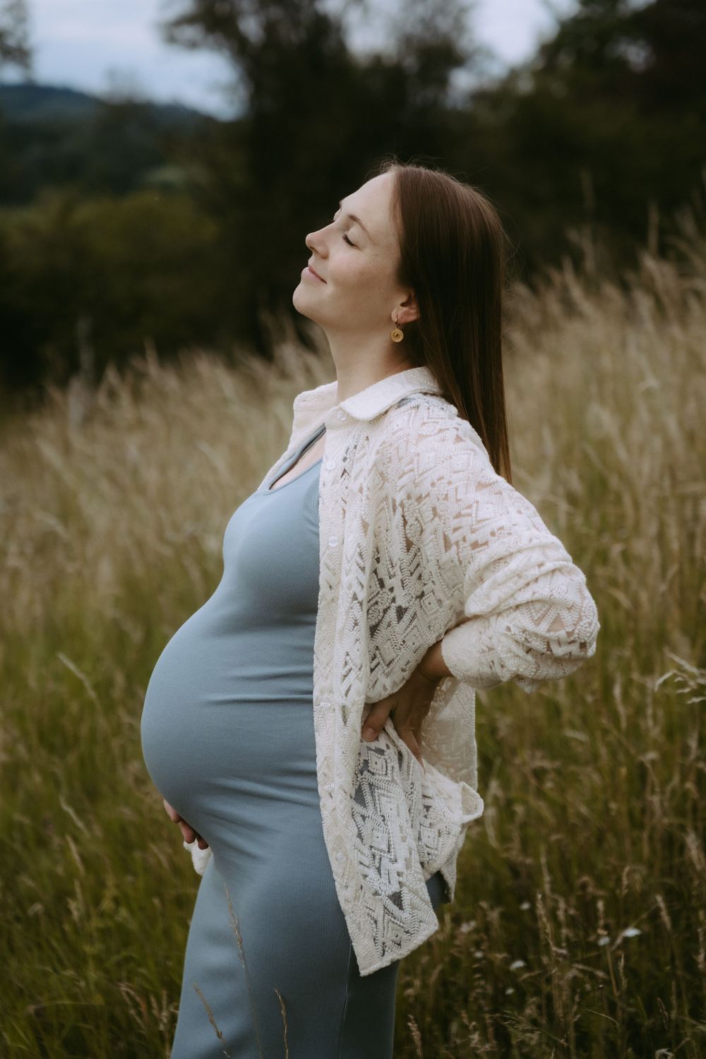 Pregnant woman in blue dress and white lace cardigan standing peacefully in tall grass field with eyes closed.