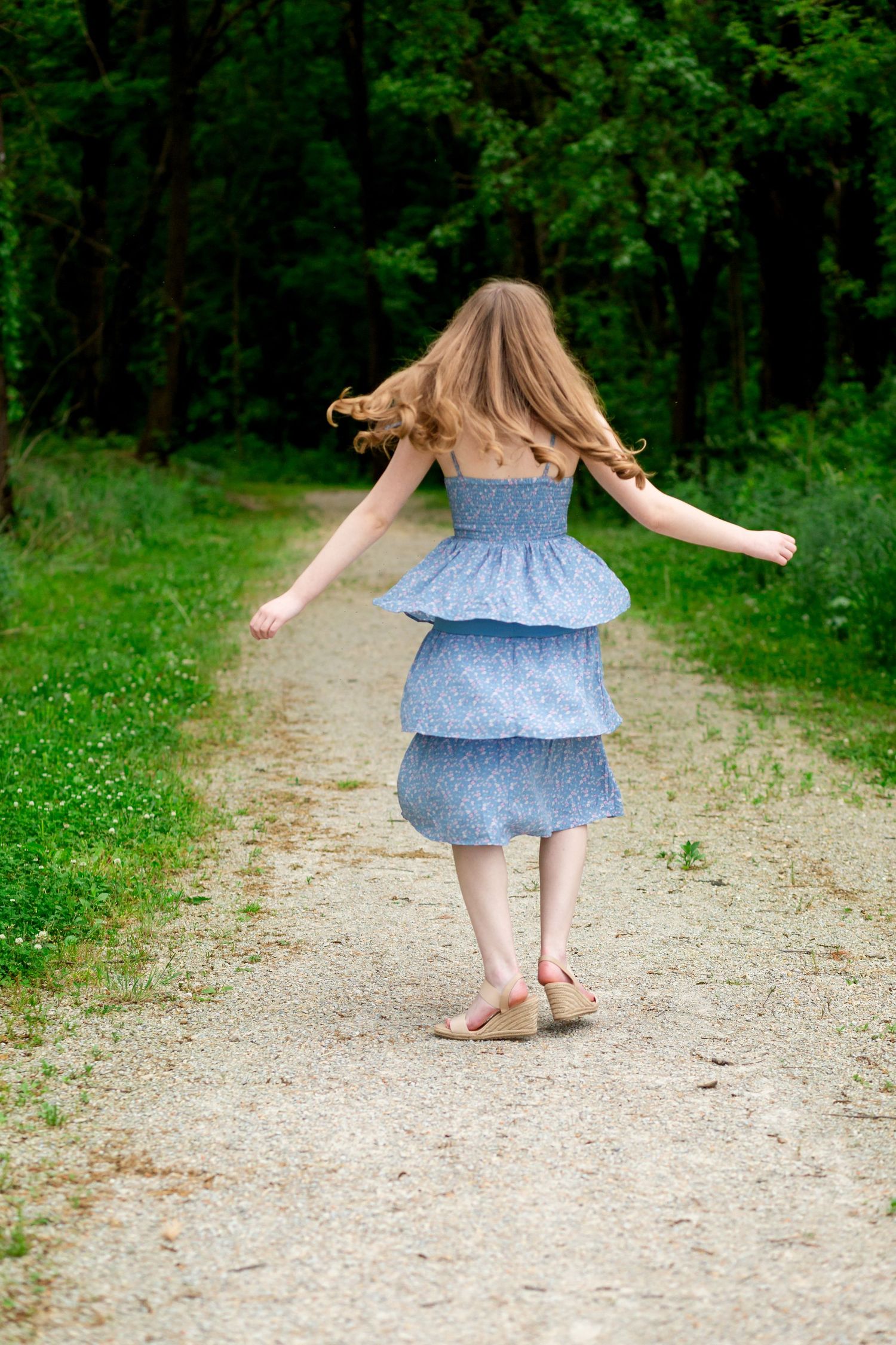 Young girl in blue tiered dress walking down a forest path with arms outstretched, long hair flowing behind her.