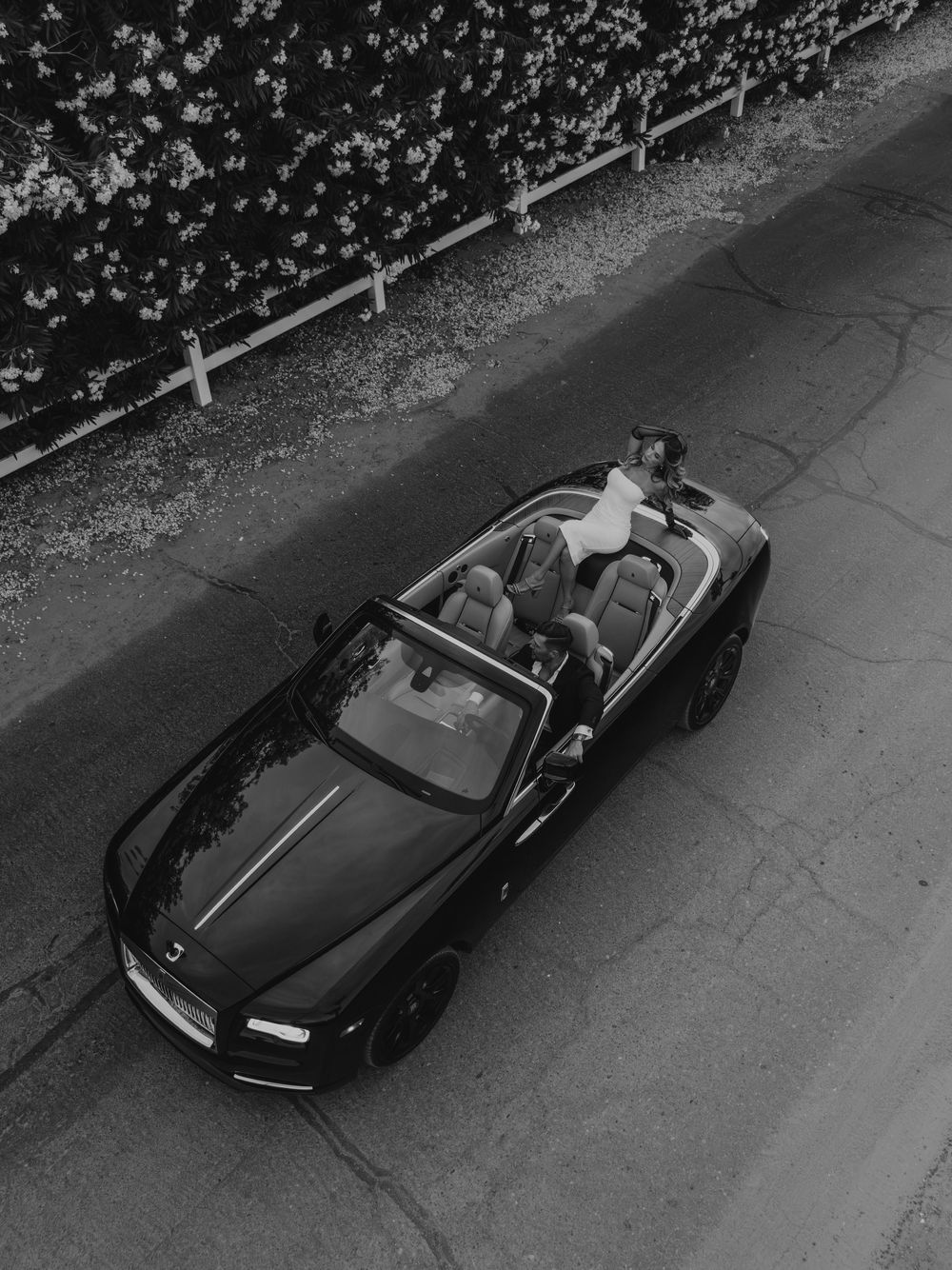 Aerial black and white photo of a luxury convertible sports car parked on a road near a hedge.