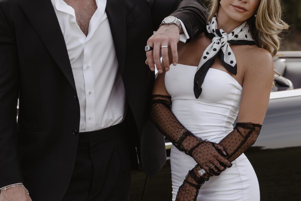 Elegant couple posing together, man in black suit, woman in white strapless dress with polka dot gloves and scarf.