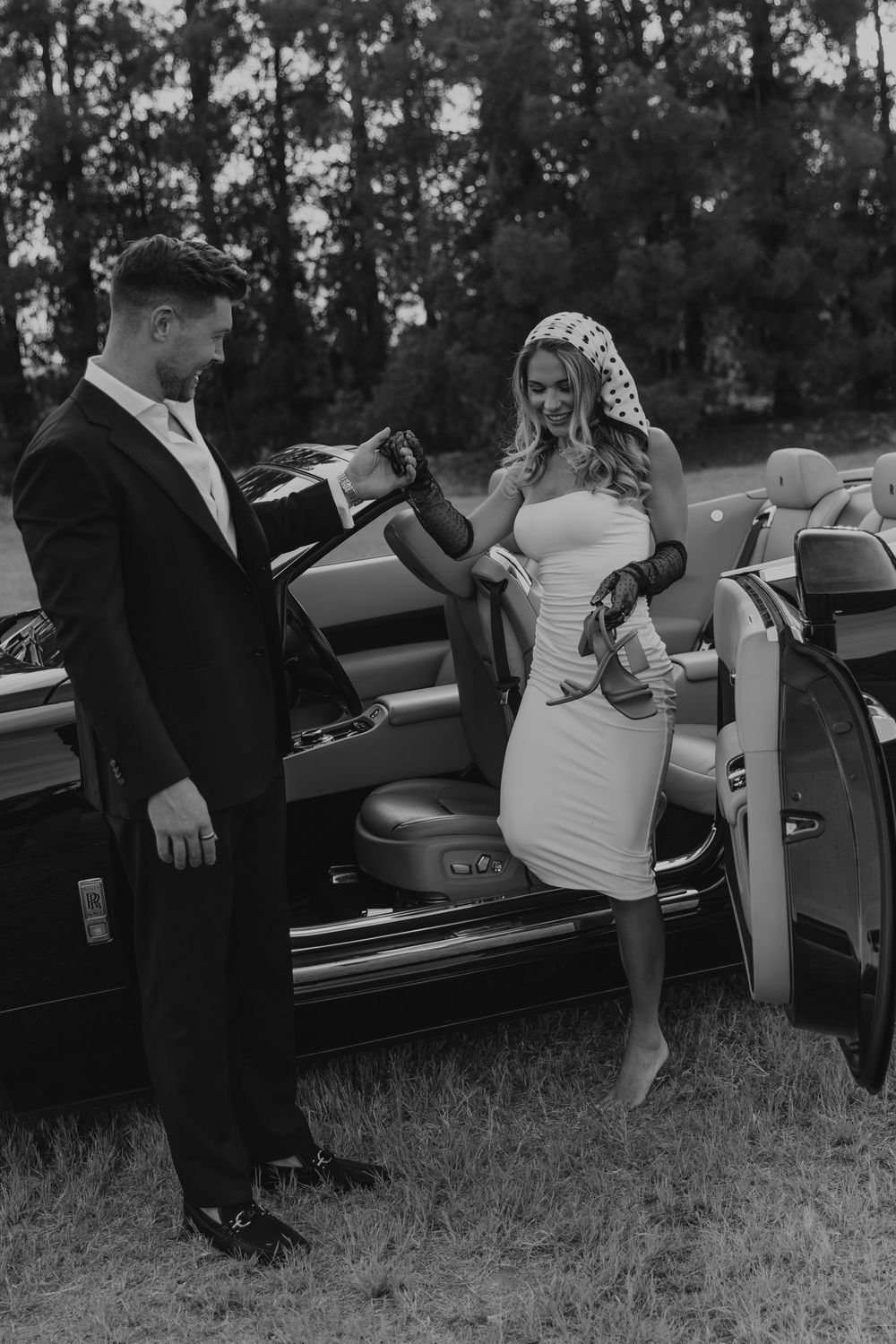 Black and white photo of a bride in white dress stepping out of a vintage convertible car assisted by groom in suit.
